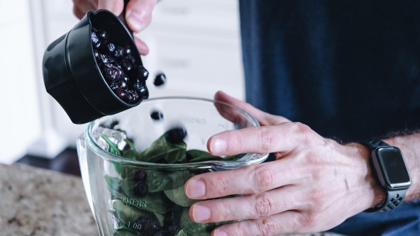 2 Hände befüllen eine Glasschüssel mit grünen Blättern und schwarzen Beeren