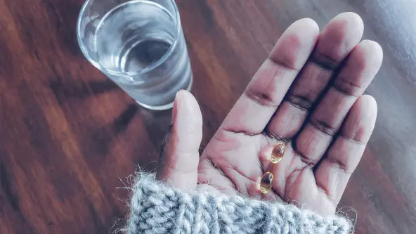 2 Tabletten liegen auf einer Hand und daneben ist ein Glas Wasser auf dem Tisch