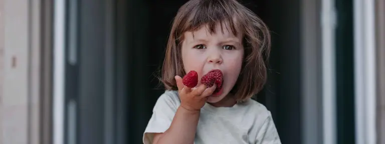 Kleines Kind steht auf einer Türschwelle und hat an ihren Fingerspitzen rote Früchte stecken