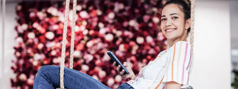Eine junge Frau sitzt mit ihrem Smartphone in einem großen Zimmer auf einer Schaukel