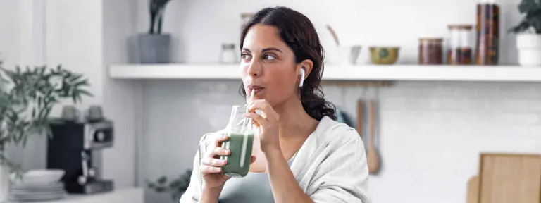 Junge, dunkelhaarige Frau in Sportoutfit und mit Airpods in den Ohren trinkt ein grünes Getränk aus einem Strohhalm