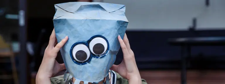Junger Mann setzt sich blaue Papiertüte auf Kopf, auf der 2 große Augen abgedruckt sind