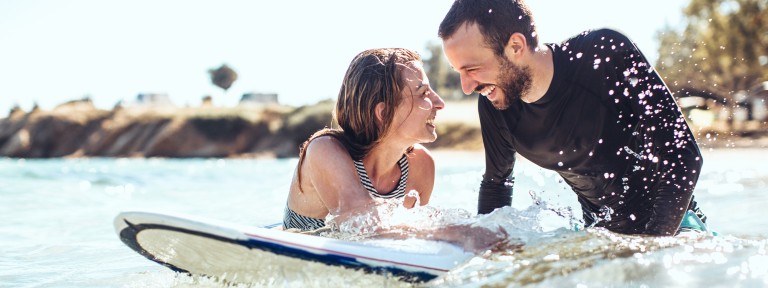 Mann und Frau liegen auf einem Surfbrett im Wasser und lachen sich dabei an