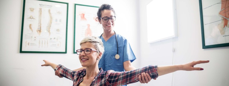 Frau mit Brille hält ihre Arme seitlich hoch und wird von Ärztin untersucht