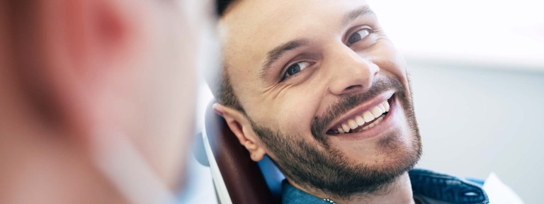 Junger Mann mit Bart lacht Zahnarzt auf Behandlungsstuhl an