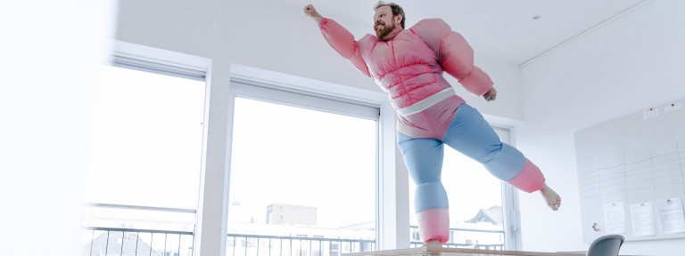 Mann in pinkem Kostüm steht als Superheld auf einem Tisch