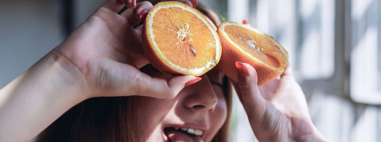 Frau hält sich 2 Hälften einer Orange an die Stirn und streckt dabei ihre Zunge raus