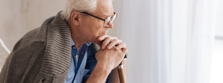 Alter Mann mit grauen Haare und Brille stützt sich sitzend auf einem Gehstock ab