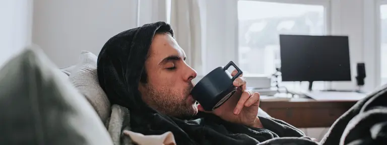 Junger Mann in einem Kapuzenpulli liegt auf einem Sofa und trinkt aus einer Tasse