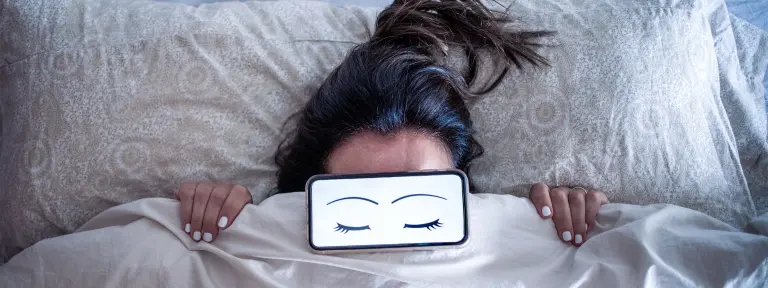 Frau hält sich Bettdecke bis unter die Augen, die mit einem Bild von geschlossenen Augen abgedeckt sind