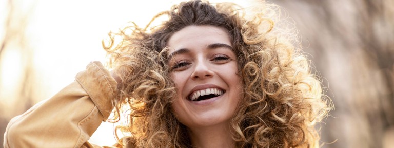Junge Frau mit lockigen Haaren blickt mit einem strahlenden Lachen in die Kamera