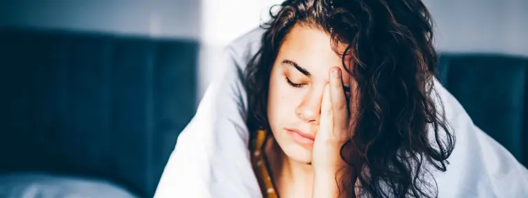 Junge Frau mit Locken sitzt mit einer Bettdecke um ihren Körper auf einem Bett und reibt sich die Stirn