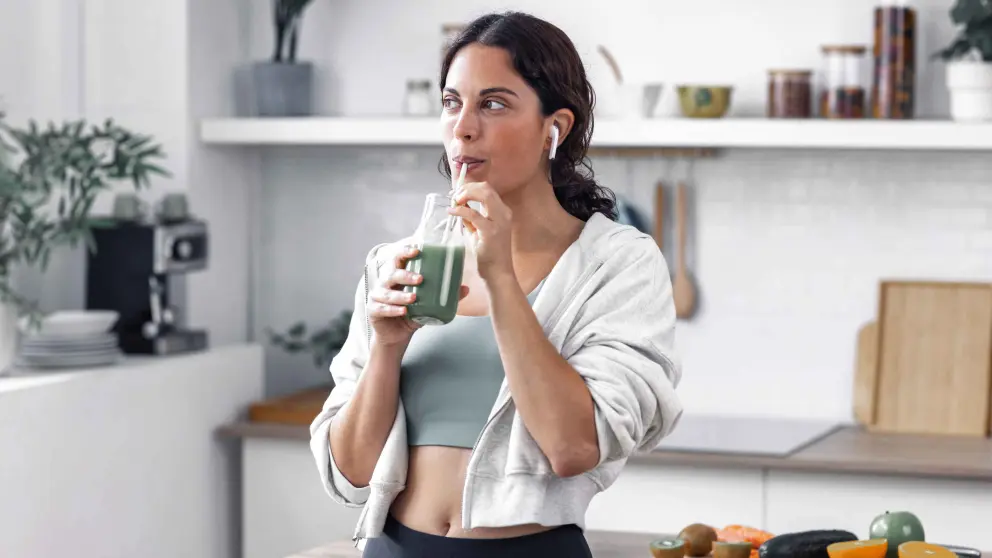 Junge, dunkelhaarige Frau in Sportoutfit und mit Airpods in den Ohren trinkt ein grünes Getränk aus einem Strohhalm