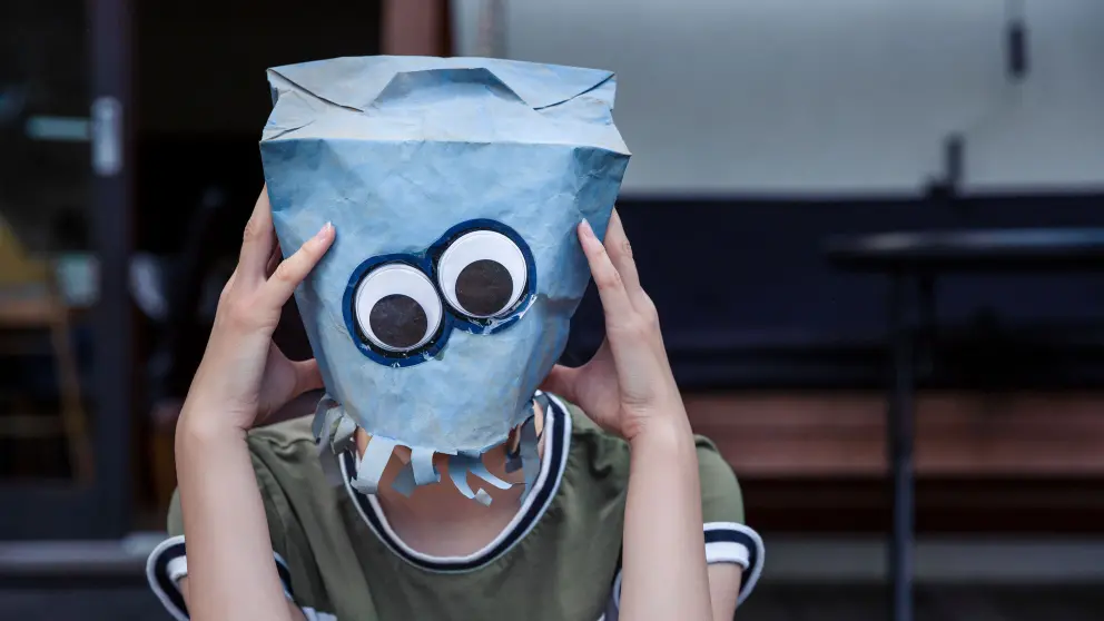 Junger Mann setzt sich blaue Papiertüte auf Kopf, auf der 2 große Augen abgedruckt sind