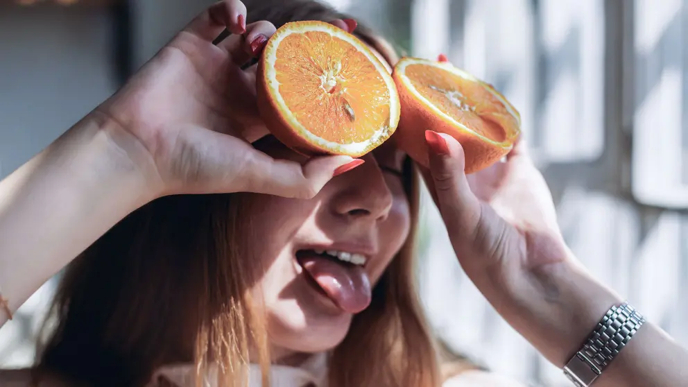 Frau hält sich 2 Hälften einer Orange an die Stirn und streckt dabei ihre Zunge raus