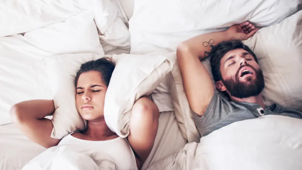 In einem Bett liegt ein schnarchender Mann und eine Frau die sich durch die Lautstärke das Kissen an die Ohren drückt