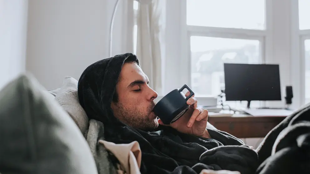 Junger Mann in einem Kapuzenpulli liegt auf einem Sofa und trinkt aus einer Tasse