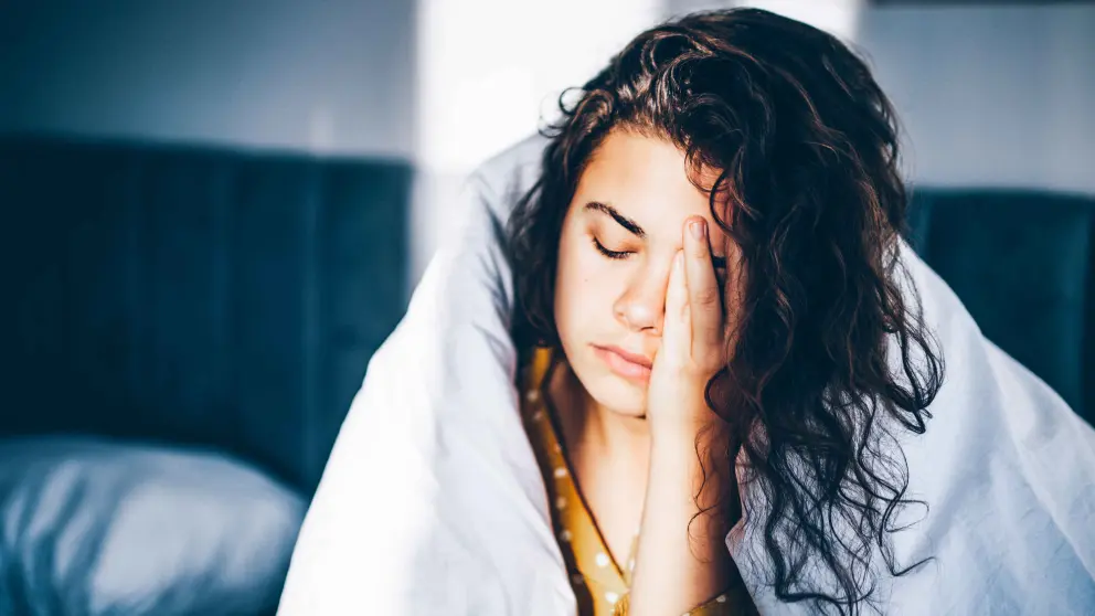 Junge Frau mit Locken sitzt mit einer Bettdecke um ihren Körper auf einem Bett und reibt sich die Stirn