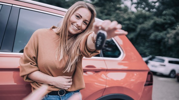 Junge Frau steht lächelnd vor ihrem Auto und hält ihren Autoschlüssel ins Bild