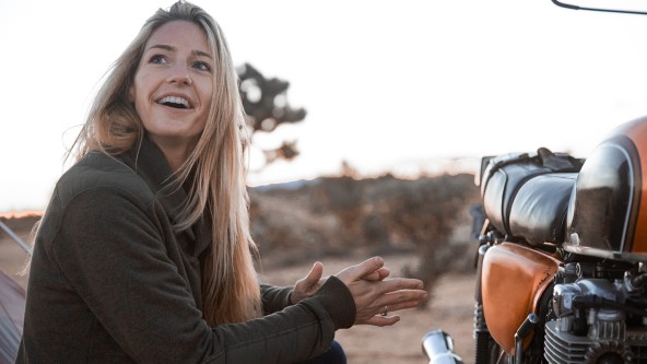 Junge blonde Frau kniet neben ihrem Motorrad und schaut erstaunt
