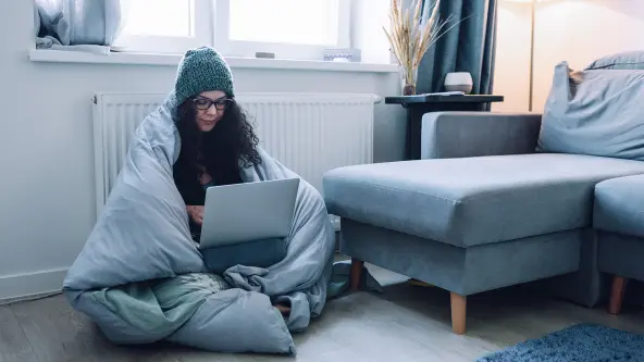 Frau sitzt mit Mütze und Decke auf Fußboden und hat Laptop auf Schoß