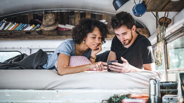 Paar liegt auf einer Matratze in einem Wohnwagen und schaut auf ein Smartphone