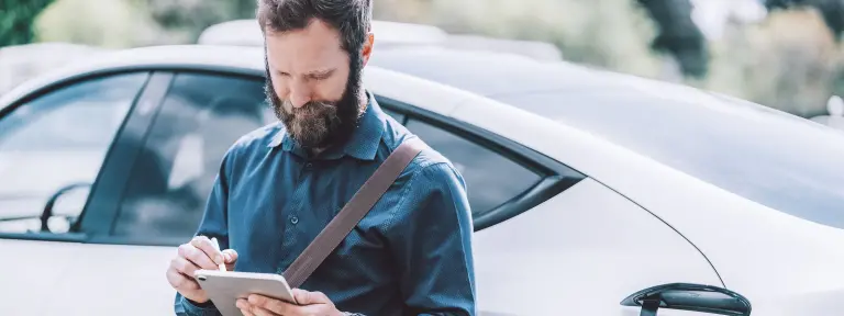 Mann lehnt an seinem weißen E-Auto und schaut auf sein Handy