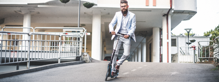 Junger Mann mit Bart und hellblauen Anzug fährt auf E-Scooter auf der Straße