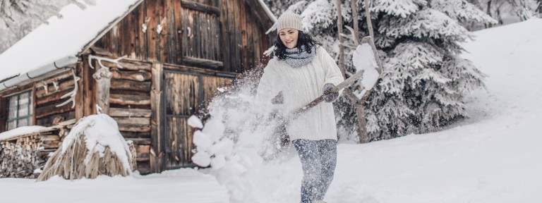 Frau schippt Schnee vor einer schneebedeckten Holzhütte