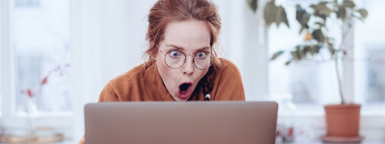 Frau sitzt vor einem Laptop und reißt ihren Mund und ihre Augen weit auf