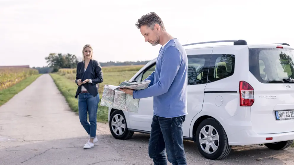Mann steht vor einem weißen Auto und schaut in eine Straßenkarte