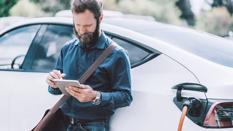 Mann lehnt an seinem weißen E-Auto und schaut auf sein Handy