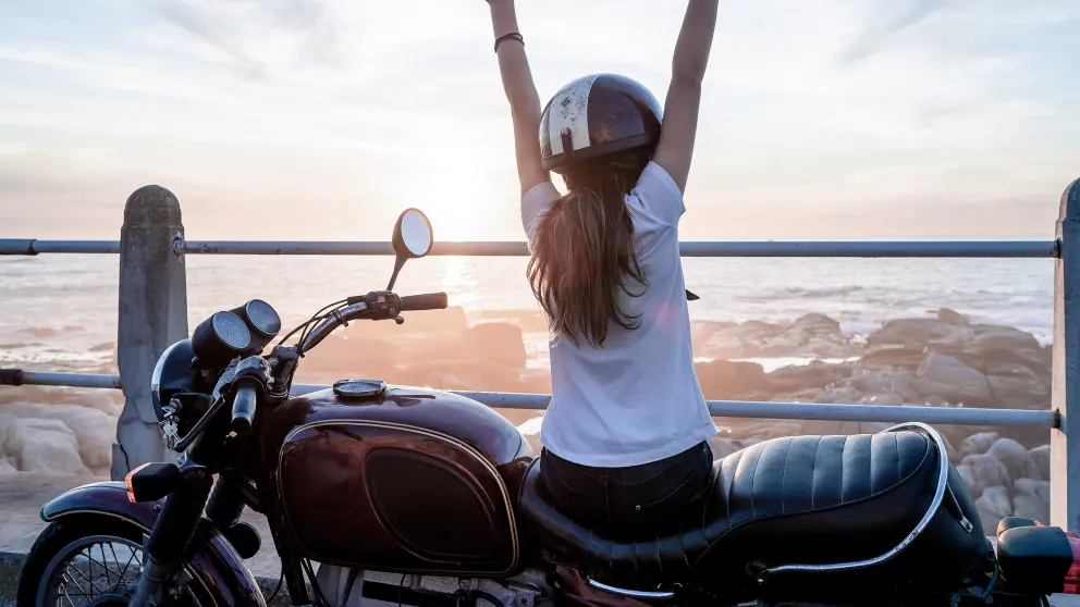 Frau auf Motorrad schaut aufs Meer und hebt ihre Hände in die Luft