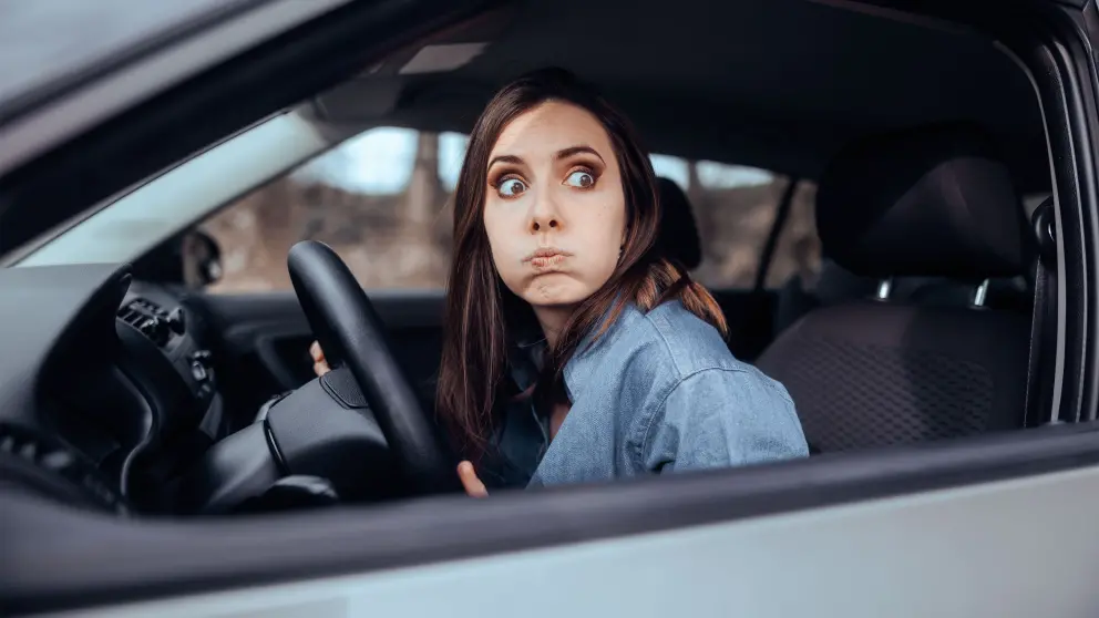 Frau mit dunklen Haaren am Steuer eines Autos schaut mit großen Augen aus dem Beifahrerfenster