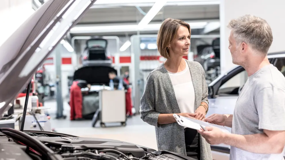 Frau und Mechaniker unterhalten sich vor geöffneter Motorhaube eines Autos
