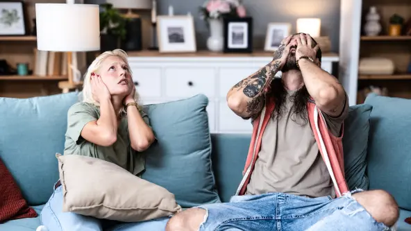 Junges Pärchen sitzt auf Sofa und hält sich die Ohren wegen lauter Musik zu. 