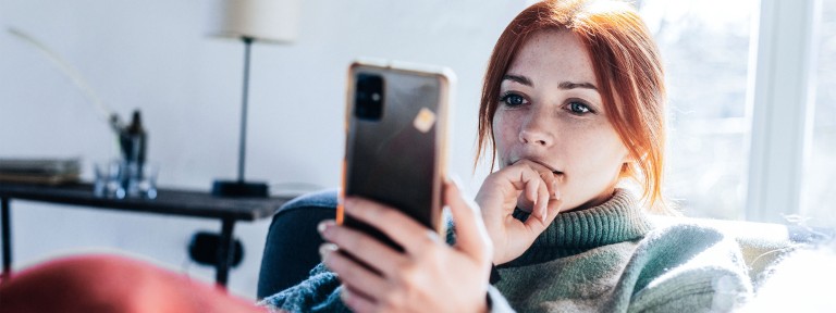 Frau mit roten Haaren sitzt auf Sofa und blickt auf ihr Smartphone
