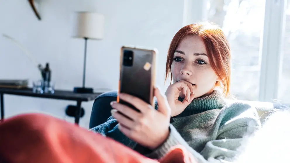 Frau mit roten Haaren sitzt auf Sofa und blickt auf ihr Smartphone