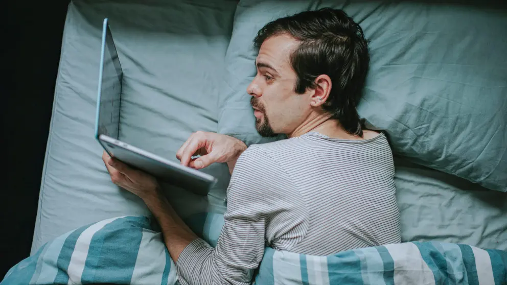 Ein junger Mann liegt mit seinem Laptop neben dem Kopf im Bett